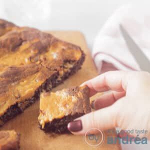 Een vierkante foto met een brownie waar een stukje van af is gesneden. Een blokje karamelbrownie wordt van de snijplank afgehaald.