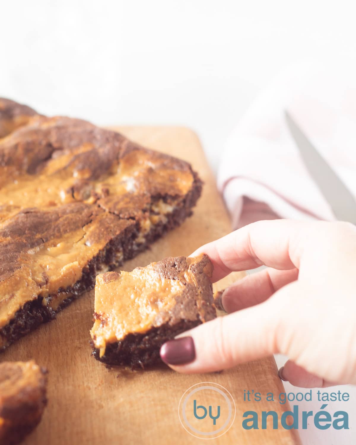 Een brownie met karamel op een houten plank. Een hand pakt een blokje er van af.