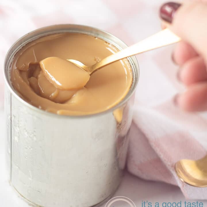 Een blikje gevuld met romige dulce de leche. Een gouden lepel haalt een beetje uit het blik. Een roze doek op de achtergrond