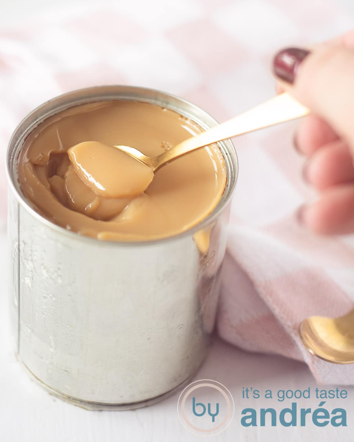 Een blikje gevuld met romige dulce de leche. Een gouden lepel haalt een beetje uit het blik. Een roze doek op de achtergrond