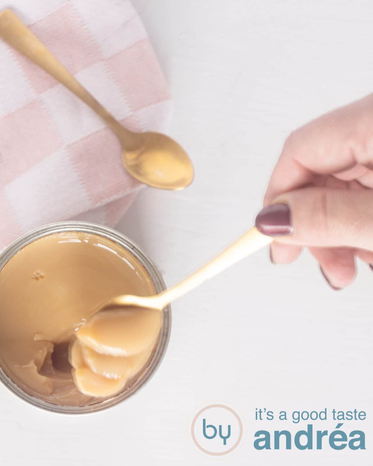 Een topfoto van zelfgemaakte dulce de leche met een lepel uit het blik gehaald. Een roze doek en gouden lepel in de top.