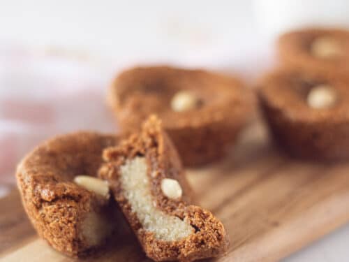 een houten bord met speculaaskoekjes gevuld met Nederlandse amandelen op een witte ondergrond. Een koekje wordt in tweeën gesneden