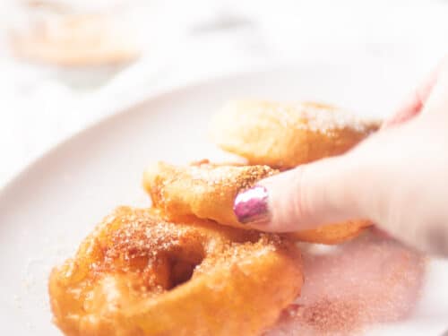 Een hand die een appelbeignet van een witte schaal met drie beignets pakt