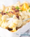 Een witte ovenschaal met daarin bloemkool gratin met spekjes. Een lepel pakt een hap van de bloemkool uit de schaal.
