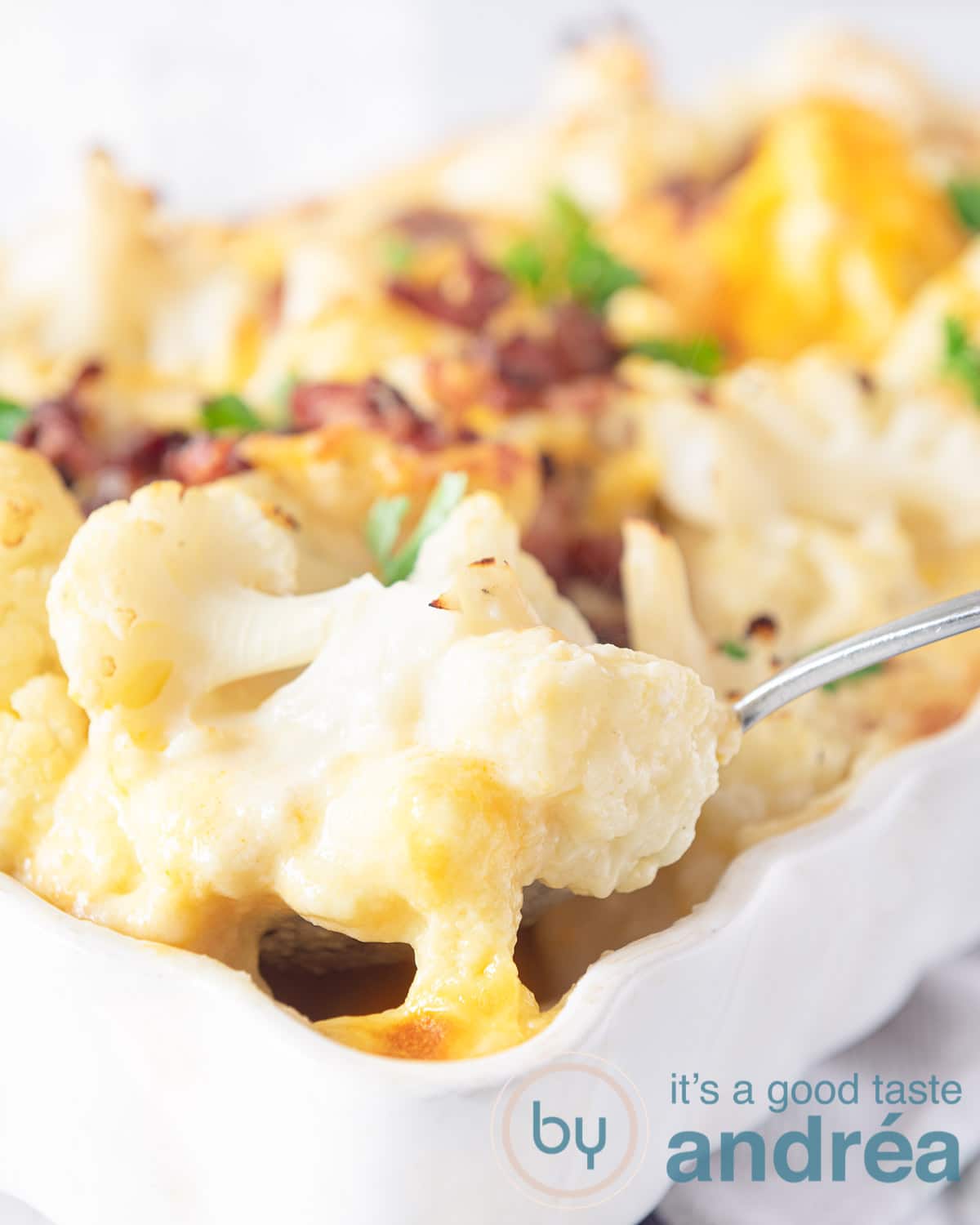 Een witte ovenschaal met daarin bloemkool gratin met spekjes. Een lepel pakt een hap van de bloemkool uit de schaal.