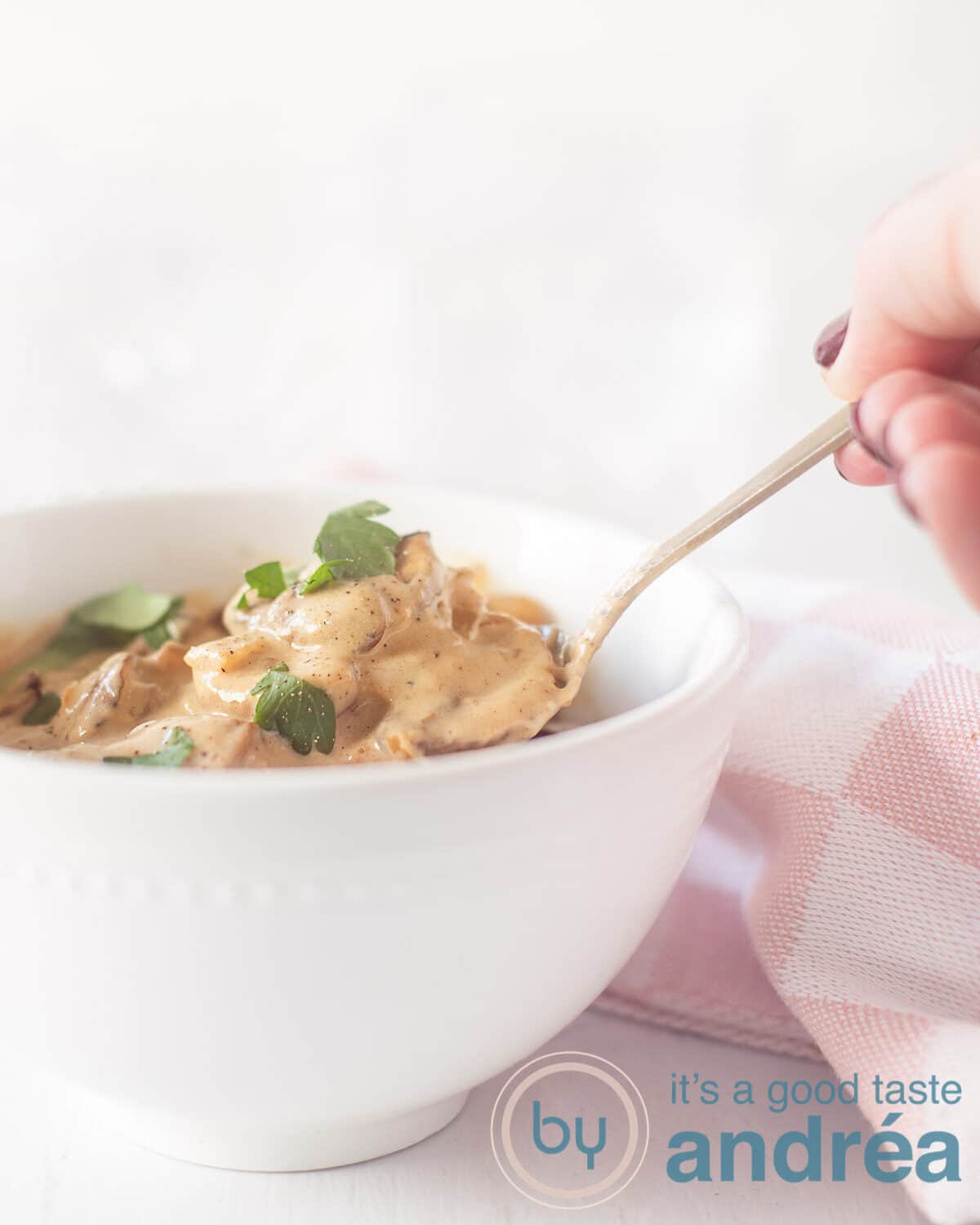 Een schaal gevuld met champignon in kookroom saus. Een gouden lepel en een hand die een schep eruit neemt. Een roze wit geblokte theedoek aan de rechterkant