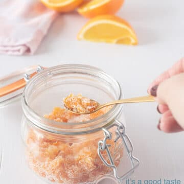 Een witte achtergrond met in de voorgrond een weckpot met gekonfijte sinaasappel. Een gouden lepel pakt een schep. Op de achtergrond een roze wit geblokte theedoek met partjes sinaasappel