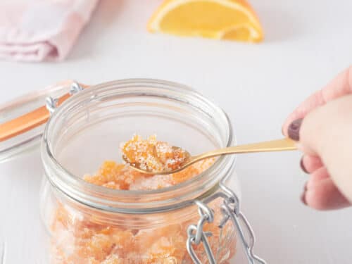 Een witte achtergrond met in de voorgrond een weckpot met gekonfijte sinaasappel. Een gouden lepel pakt een schep. Op de achtergrond een roze wit geblokte theedoek met partjes sinaasappel