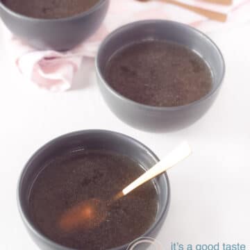 Een witte ondergrond daarop drie grijze kommen gevuld met bouillon van kip. In de voorste kom staat een gouden lepel. In de achtergrond een roze wit geblokte theedoek met daarop twee gouden lepels