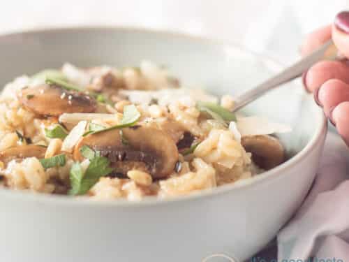 Een blauw schaaltje met risotto, kastanjechampignons en kruiden. Een hand pakt een lepel. Een witte achtergrond