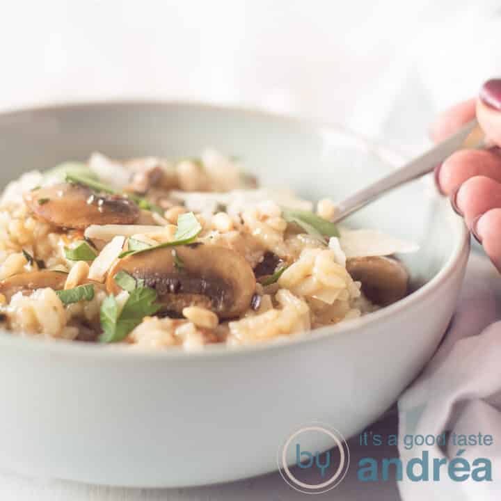 Een blauw schaaltje met risotto, kastanjechampignons en kruiden. Een hand pakt een lepel. Een witte achtergrond