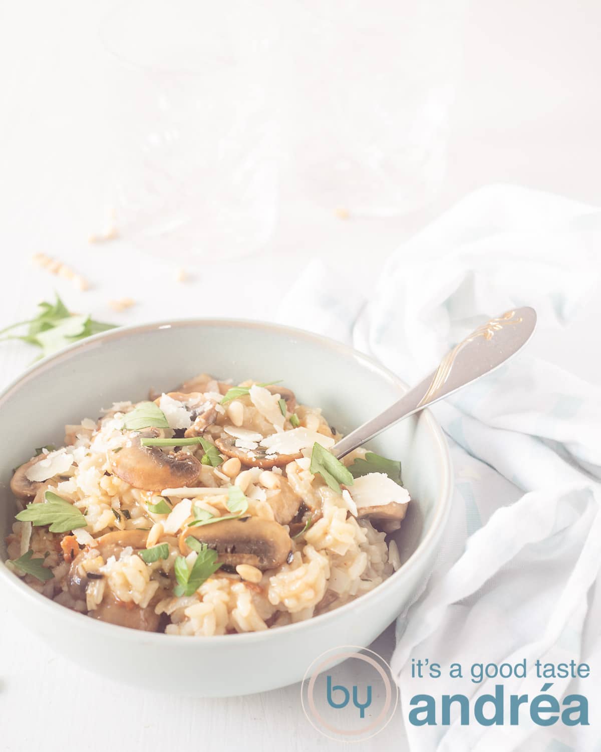 Een blauwgroen schaaltje linksonder met een lepel, gevuld met risotto, kastanjechampignon, peterselie en pijnboompitten. Op een witte achtergrond. Een witte met blauwe blokjes theedoek rechts.