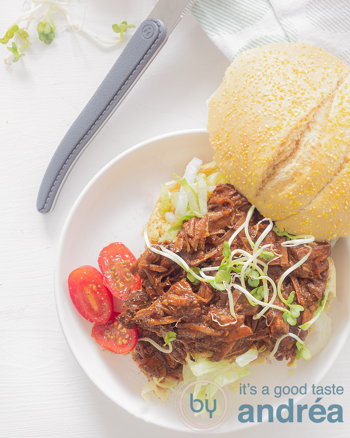 Een foto van bovenaf van een broodje met daarop shredded beef, tomaten en cress. Een witte achtergrond. Bovenin een mes met een grijs handvat