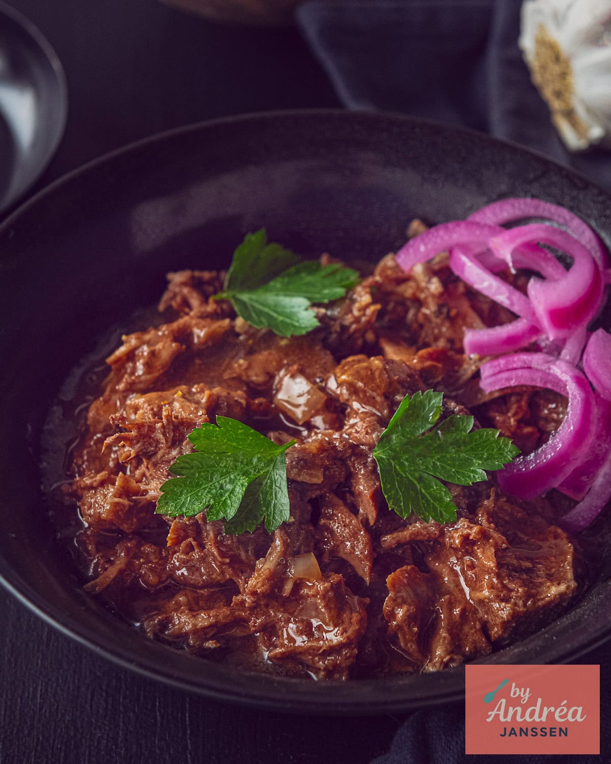 A bowl filled with slow cooked pulled beef, topped with parsley and sweet and sour red onions.