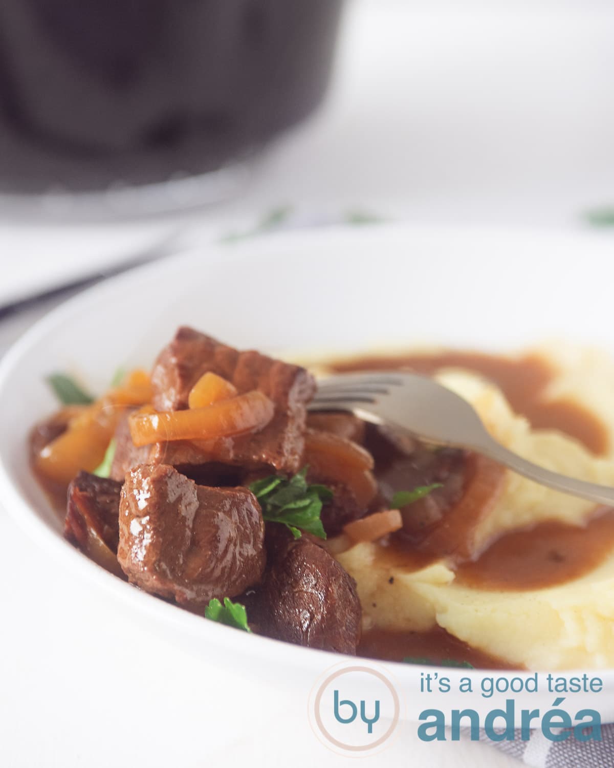 Een half wit diep bord met aardappelpuree, hachee met uien en heerlijke jus. Een gouden vork prikt in een stukje vlees. De slowcooker op de achtergrond