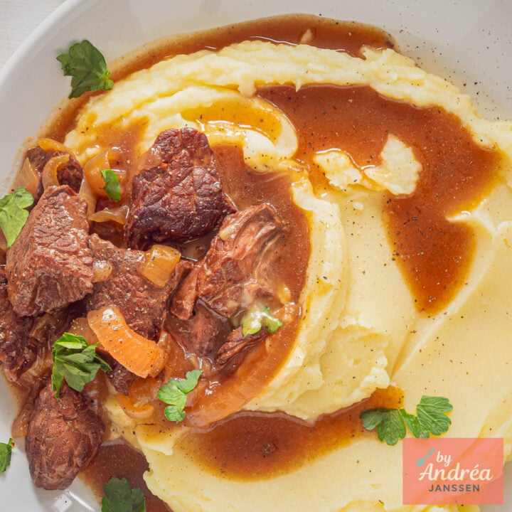 Detail van stukken rundvlees en uien in bruine jus naast aardappelpuree met peterselie, van bovenaf gefotografeerd.