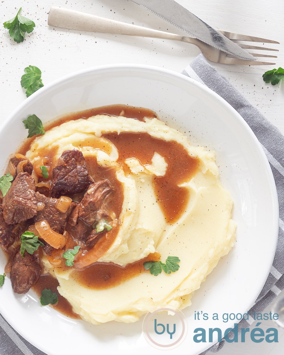 Een foto van bovenaf van een wit bord gevuld met aardappelpuree, stukjes vlees (hachee) en jus. Peterselie eromheen. Op een witte ondergrond. Erboven een gouden vork en een mes.