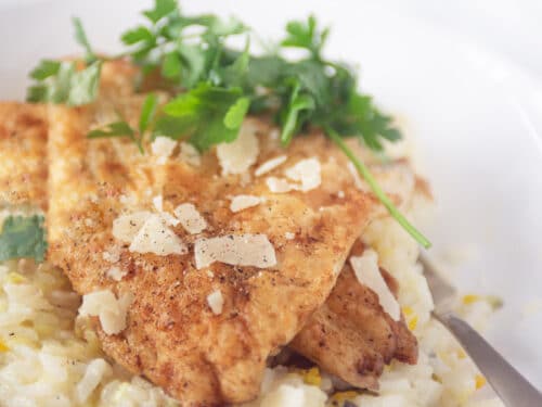 Foto van een wit bord met een gouden lepel. Het bord is gevuld met risotto met courgette en kaas. Daarboven op liggen twee moten gebakken kabeljauw