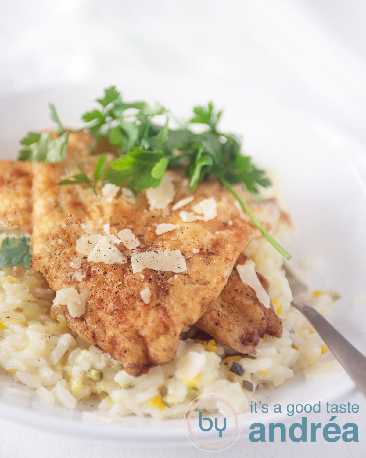 Foto van een wit bord met een gouden lepel. Het bord is gevuld met risotto met courgette en kaas. Daarboven op liggen twee moten gebakken kabeljauw