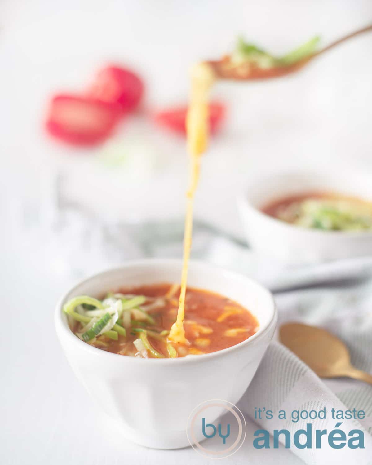 Twee kommen tomatensoep (een vooraan in het midden) met prei en kaas. Een lepel neemt een hap en gesmolten kaas hangt tussen de soep en de lepel. Een groen witte theedoek ligt tussen de kommen met daarop twee gouden lepels. Tomaten in blokjes op de achtergrond