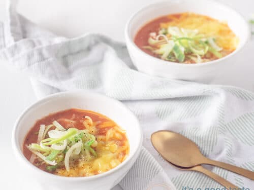 Een vierkante foto van een witte ondergrond met twee witte kommen gevuld met tomatensoep. Met een topping van prei en kaas. Twee gouden lepels linksonderin