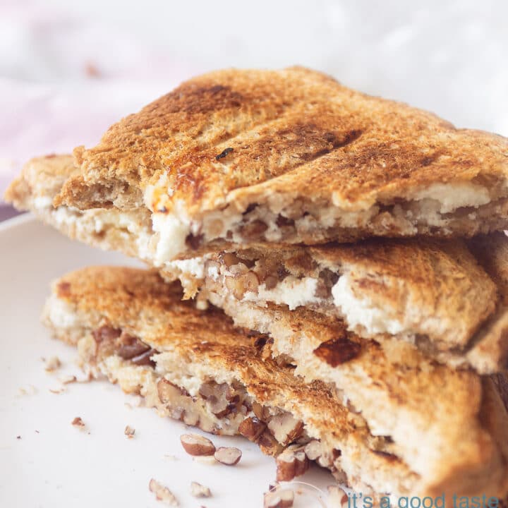 Een hoogtefoto met een macro van drie halve sneetjes tosti gevuld met geitenkaas, walnoten. In de achtergrond een witte ondergrond en een roze wit geblokte theedoek