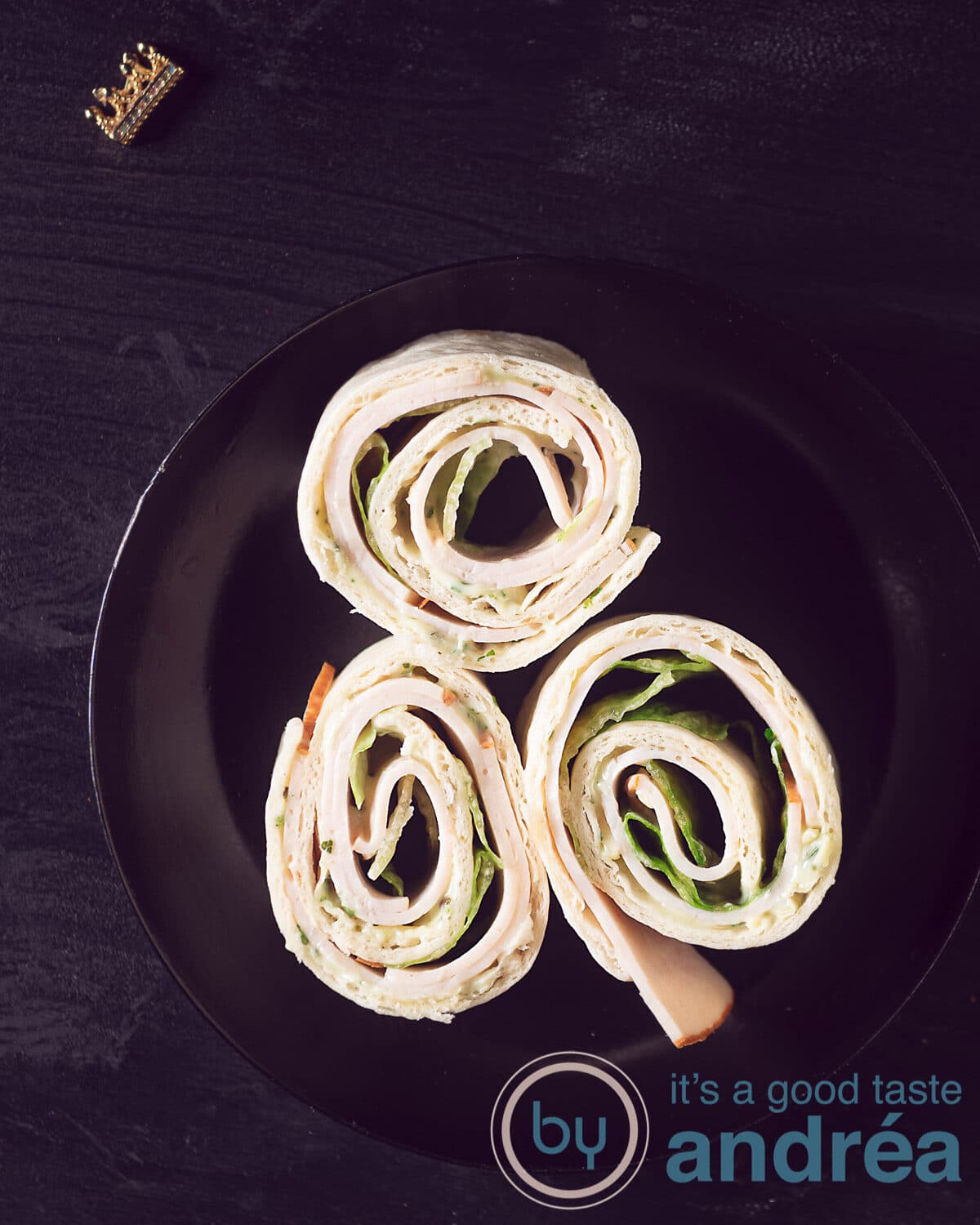 Een zwart bord met drie opgerolde tortilla rolletjes met kipfilet, ijsbergsla en heksenkaas. Alles op een zwart bord dat staat op een zwarte ondergrond