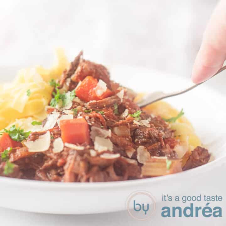 Een vierkante foto met een wit bord gevuld met pappardelle en beef ragout met wortel en Parmezaanse kaas. Een vork prikt erin.