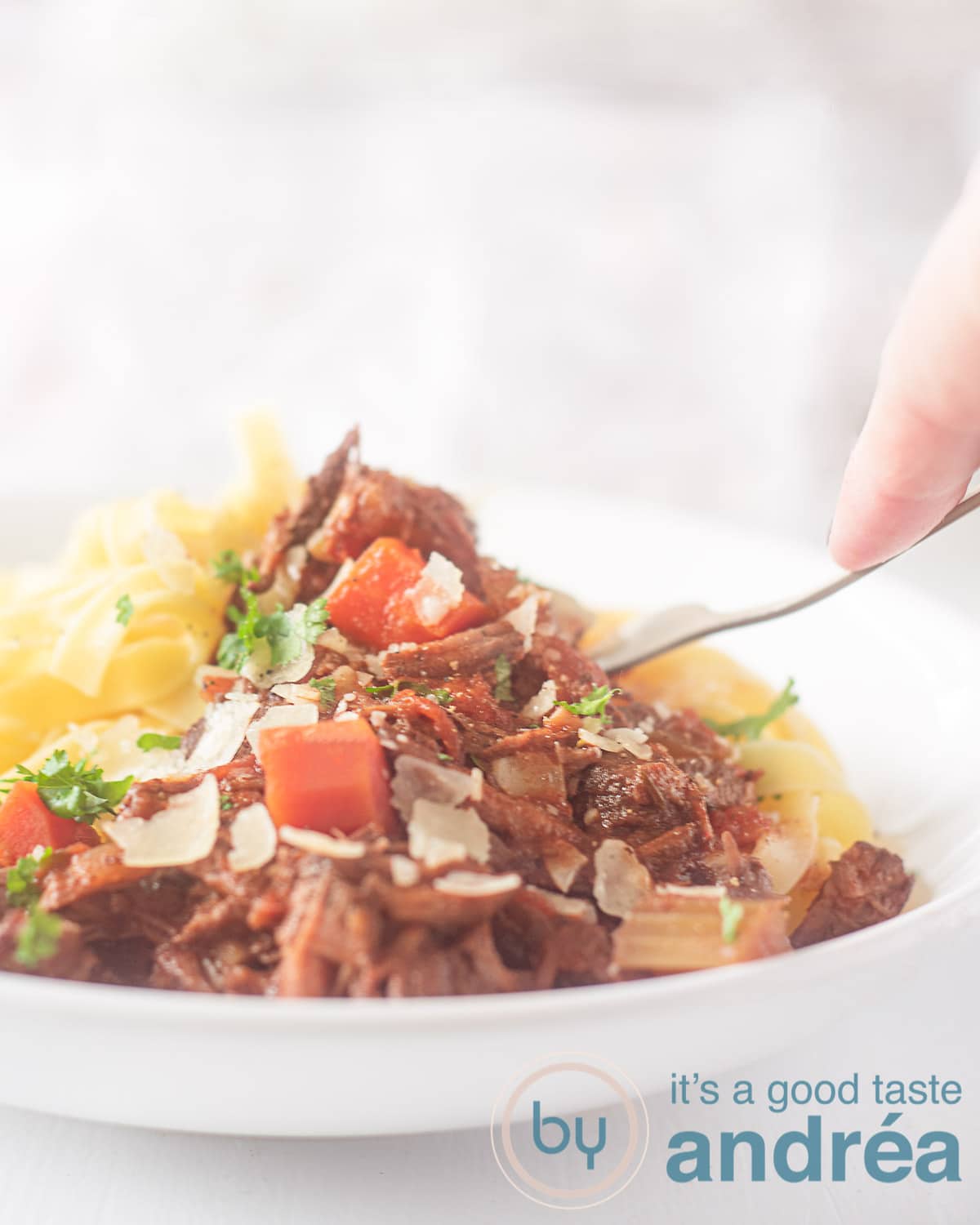 Een wit bord gevuld met Beef Ragu met wortel en Parmezaanse kaas en papardelle. Op een witte ondergrond. Een vork prikt in een stukje vlees