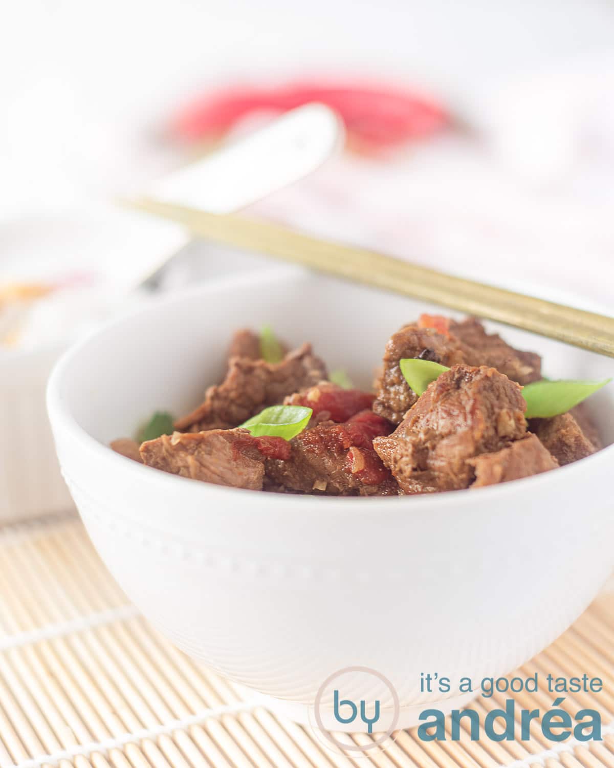 Een witte kom gevuld met daging smoor gegarneerd met pepers en lente ui. Twee chopsticks erbovenop. Het kommetje staat op een rieten matje.