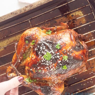 Een foto van bovenaf van een kip op een rooster in een bakblik. Heerlijk bruin met sesamzaad en peterselie. Een hand pakt de poot.
