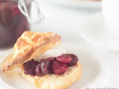 Een vierkante foto met een scone gevuld met clotted cream en cranberry compote. Een witte achtergrond.