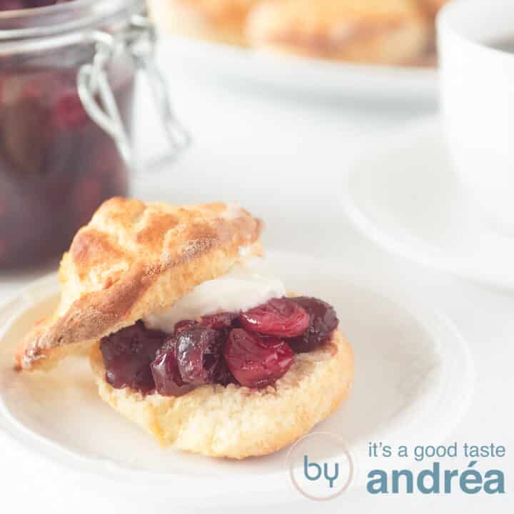 Een vierkante foto met een scone gevuld met clotted cream en cranberry compote. Een witte achtergrond.