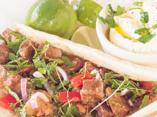 Een wit bord met een tortilla gevuld met Mexicaans stoofvlees, tomaat en ui. Op de achtergrond limoen en een kommetje met crème fraîche