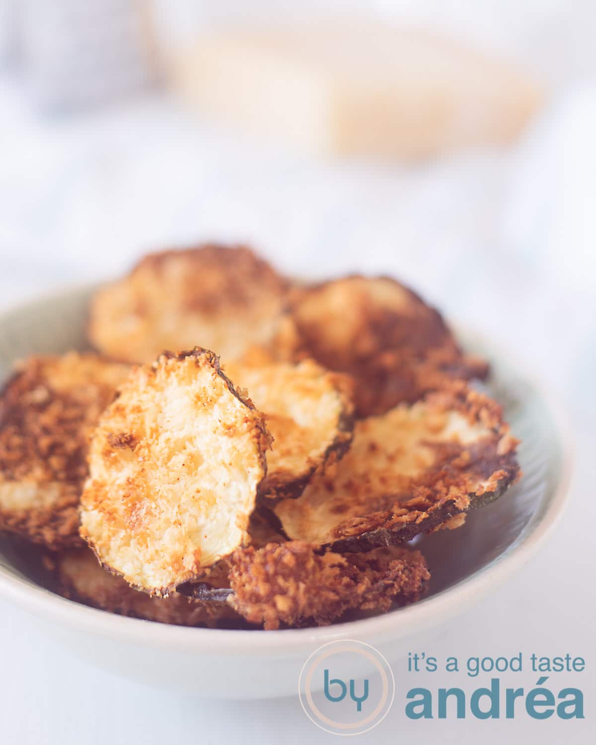 Een bakje vol met chips van courgette gegratineerd met parmezaanse kaas. Op de achtergrond ligt Parmezaanse kaas en een rasp