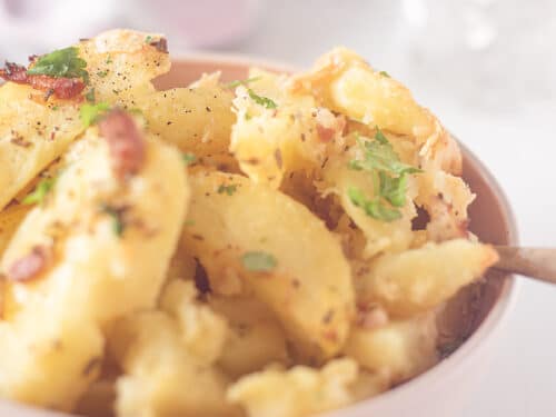 Een roze schaaltje met geroosterde knoflook aardappelen met kruiden. Twee glazen en een theedoek