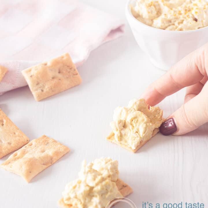 A hand grabbing a toast with chicken curry salad. A second piece of toast lies underneath. In the top left corner a pink white striped cloth and in the top right the rest of the salad
