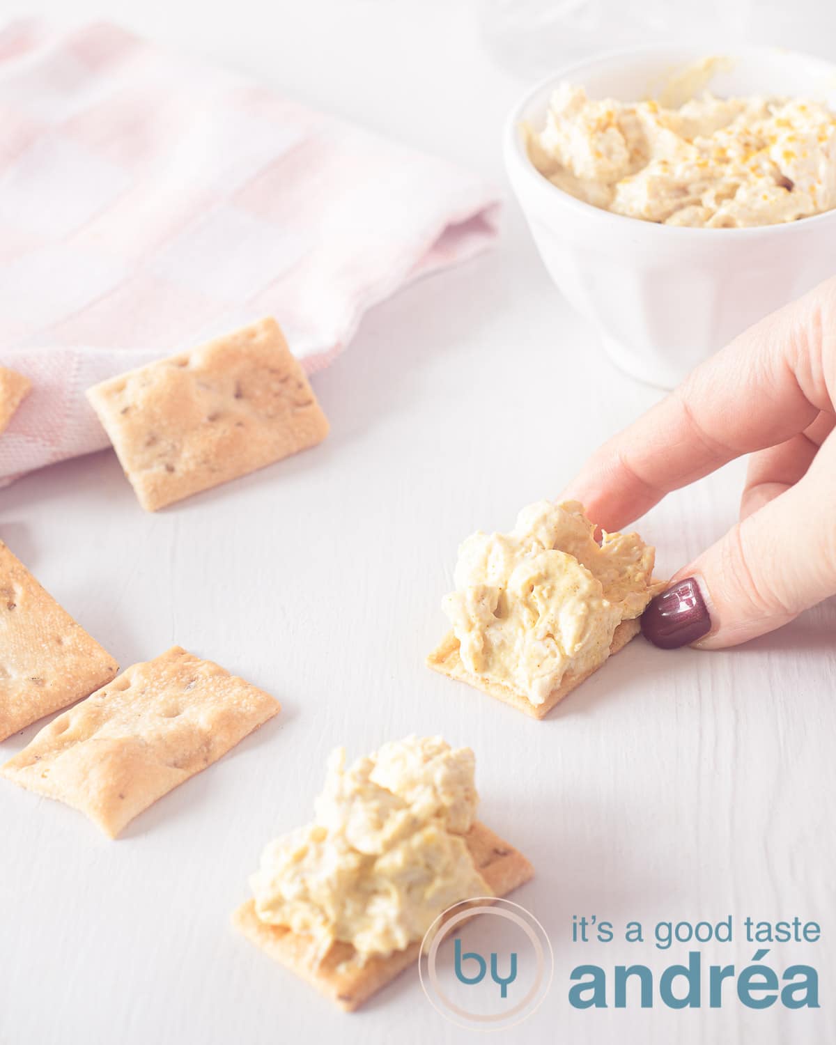 A hand grabbing a toast with chicken curry salad. A second piece of toast lies underneath. In the top left corner a pink white striped cloth and in the top right the rest of the salad