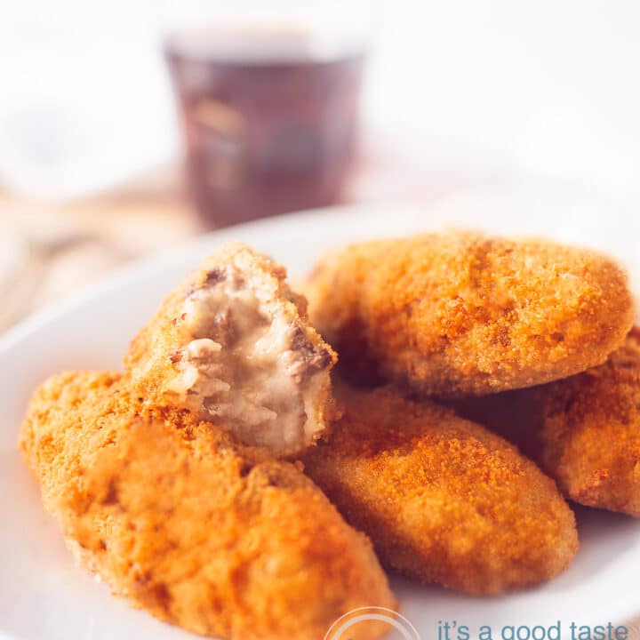 Een witte schaal met vier kroketejes. De bovenste kroket is gehalveerd, zodat je de binnenzijde kunt zien. Op de achtergrond een glas cola.