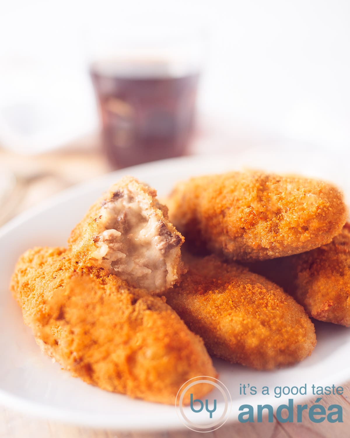 Een witte schaal met vier kroketejes. De bovenste kroket is gehalveerd, zodat je de binnenzijde kunt zien. Op de achtergrond een glas cola.