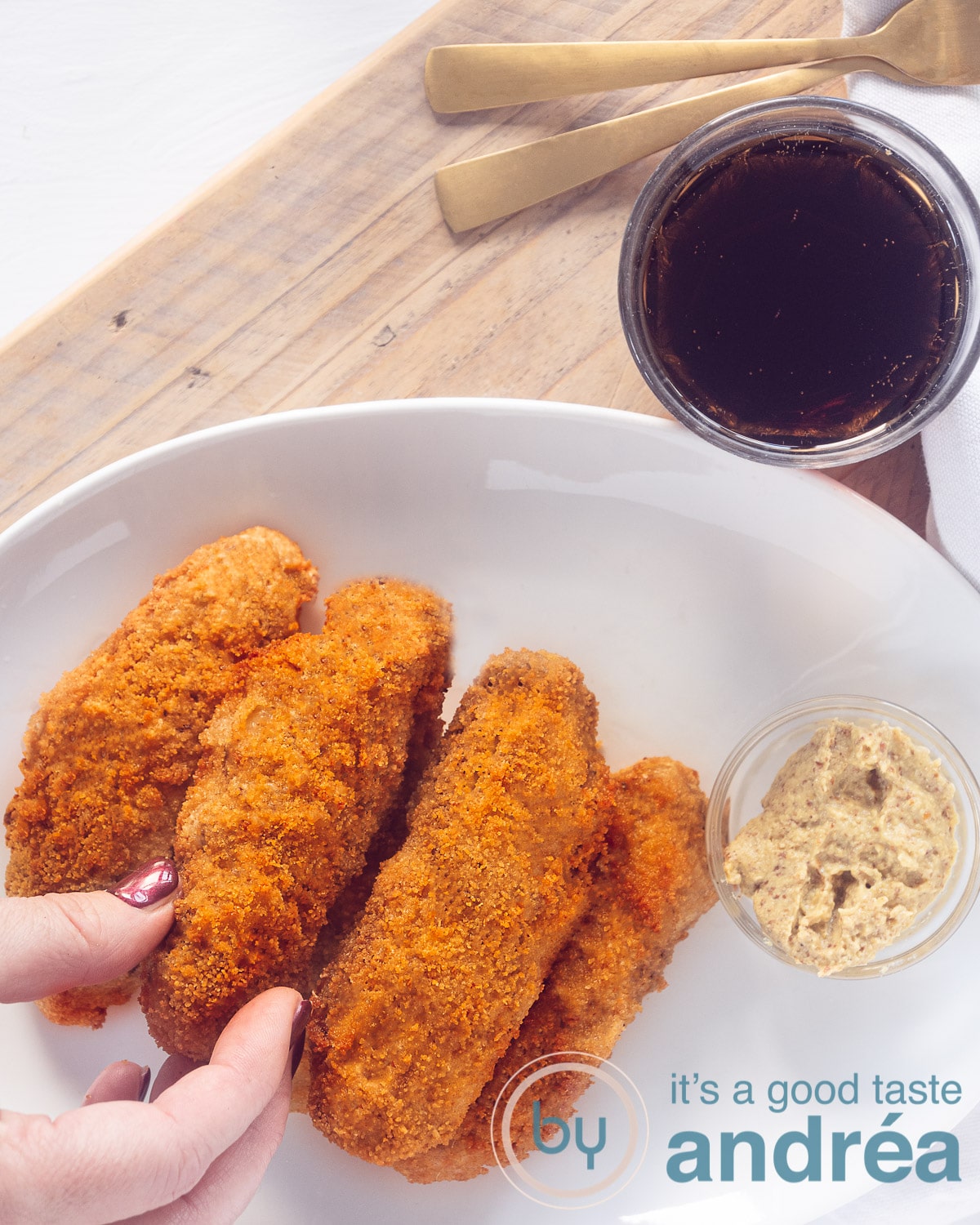 Een hand die een kroket van een witte schaal pakt (met vijf kroketten) op een houten plank. Een glas cola staat er boven