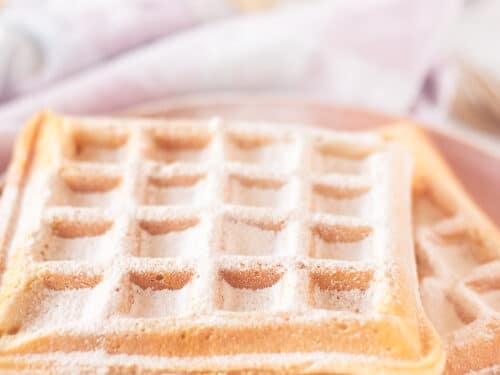 Een roze bord met daarop twee wafels bestrooid met poedersuiker. in de achtergrond een roze wit geblokte theedoek