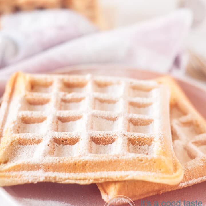 Een roze bord met daarop twee wafels bestrooid met poedersuiker. in de achtergrond een roze wit geblokte theedoek