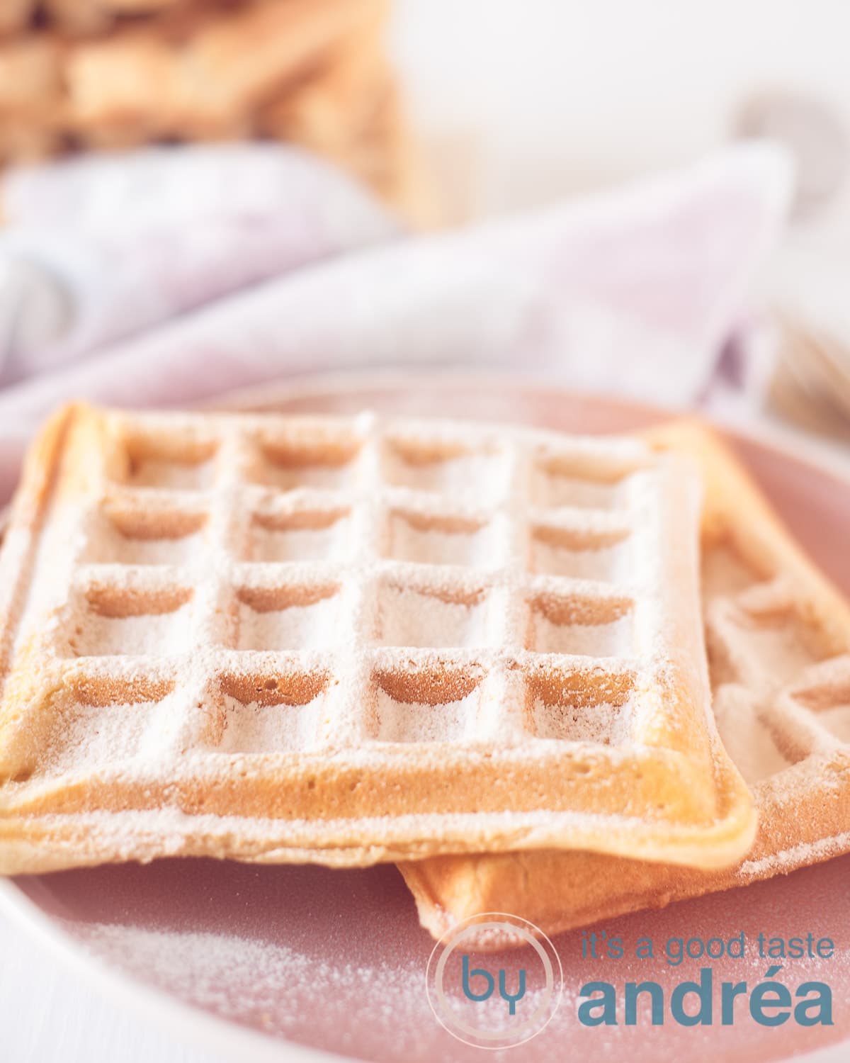 Een roze bord met daarop twee wafels bestrooid met poedersuiker. in de achtergrond een roze wit geblokte theedoek