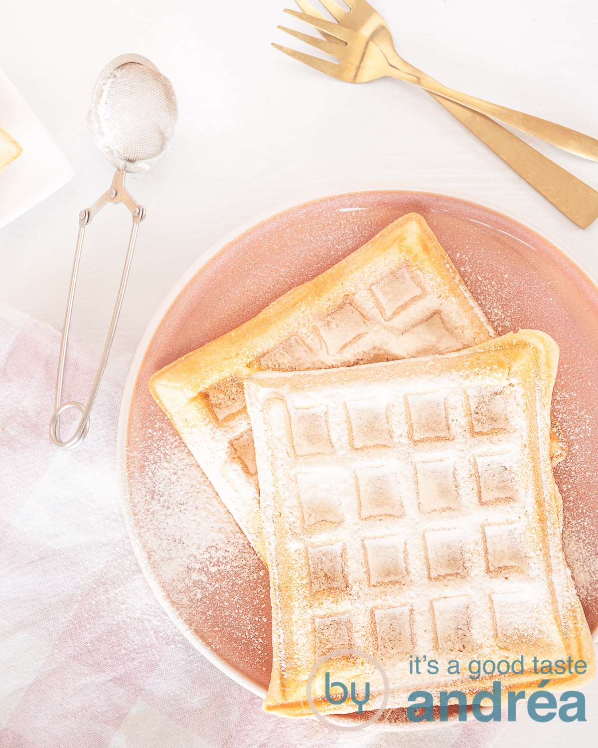 Een foto van bovenaf van ouderwetse wafels op een roze bord, bestrooid met poedersuiker. In de linker onderhoek een roze witte theedoek. Een zeef met poedersuiker en twee gouden vorken. Een witte ondergrond.