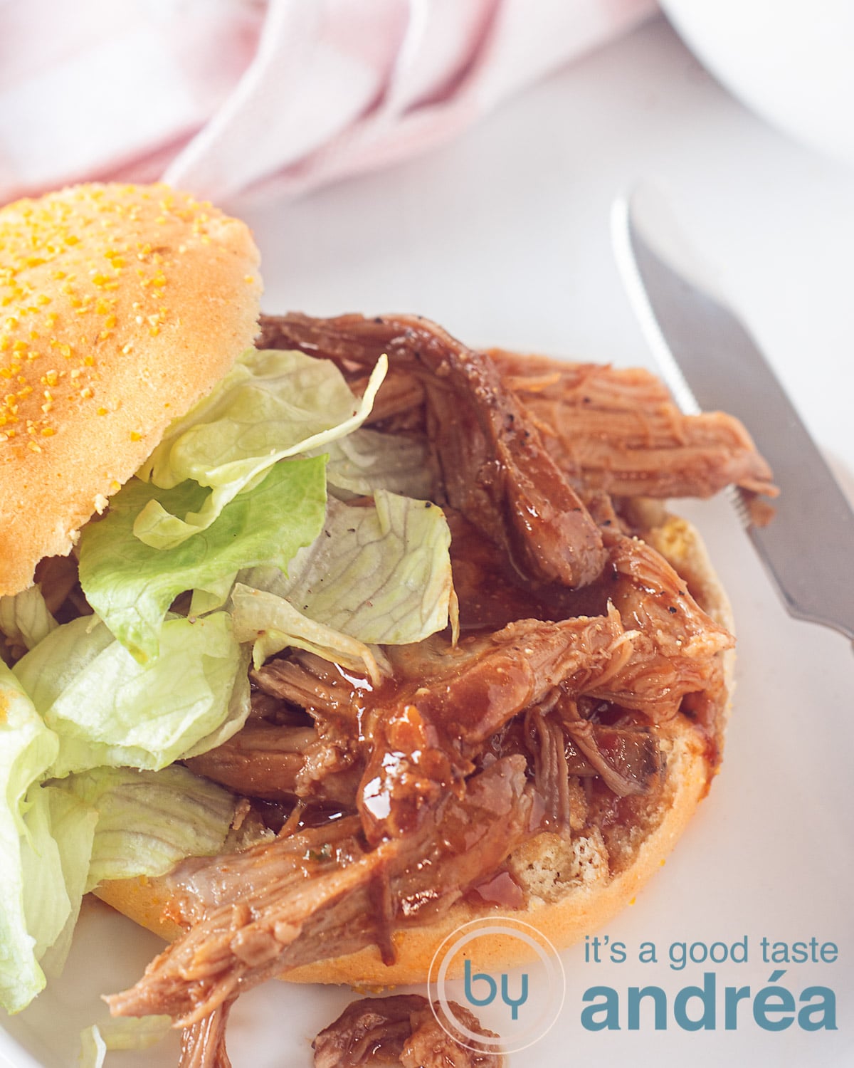 Een witte ondergrond, daarop een broodje met BBQ pulled pork en een salade. Een mes er rechts naast, een witte ondergrond