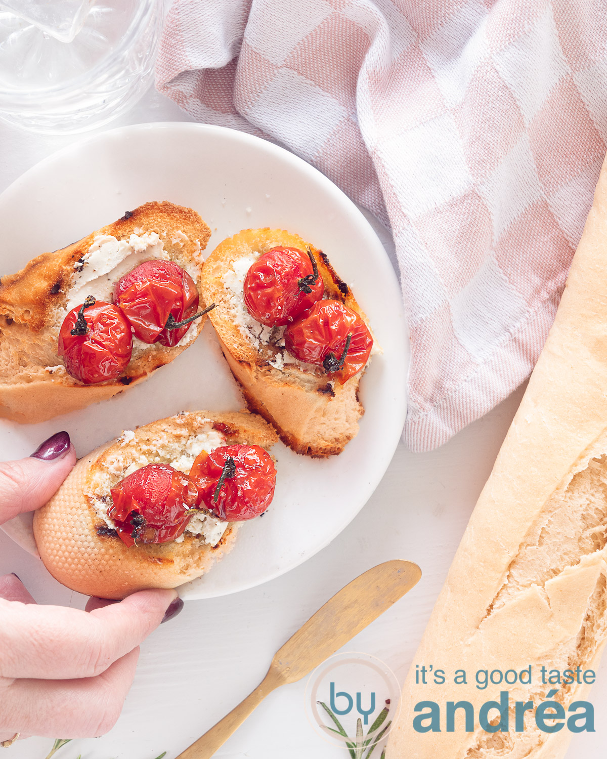 Een hoogtefoto van bovenaf. Linksbovenin een wit bord met drie sneetjes bruschetta geitenkaas met gestoofde tomaten. Een hand pakt er een. Rechts een stokbrood en een wit roze theedoek.