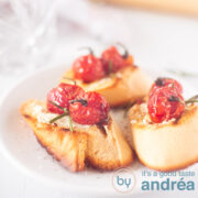 Een vierkante foto met drie stukjes gegrild stokbrood, geitenkaas en gestofode tomaten op een witte ondergrond
