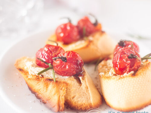 Een vierkante foto met drie stukjes gegrild stokbrood, geitenkaas en gestofode tomaten op een witte ondergrond