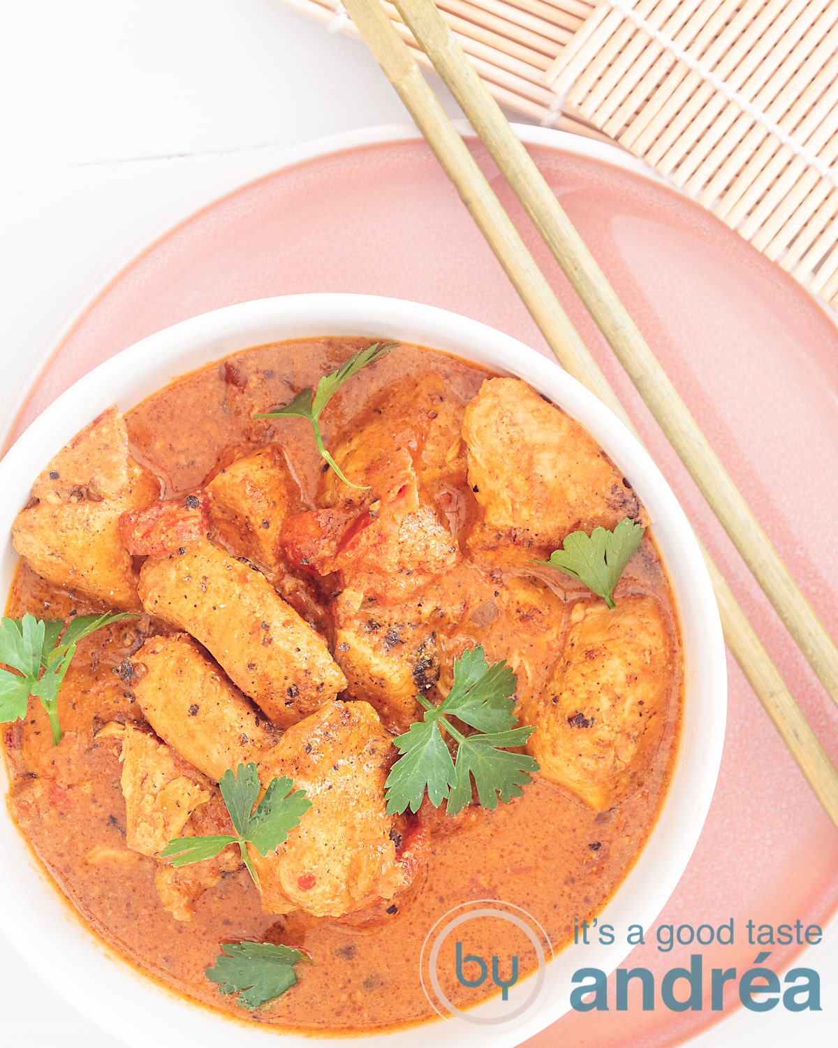 Een foto van bovenaf van kip met boterige curry saus op een roze bord. Twee chopsticks liggen er rechts naast